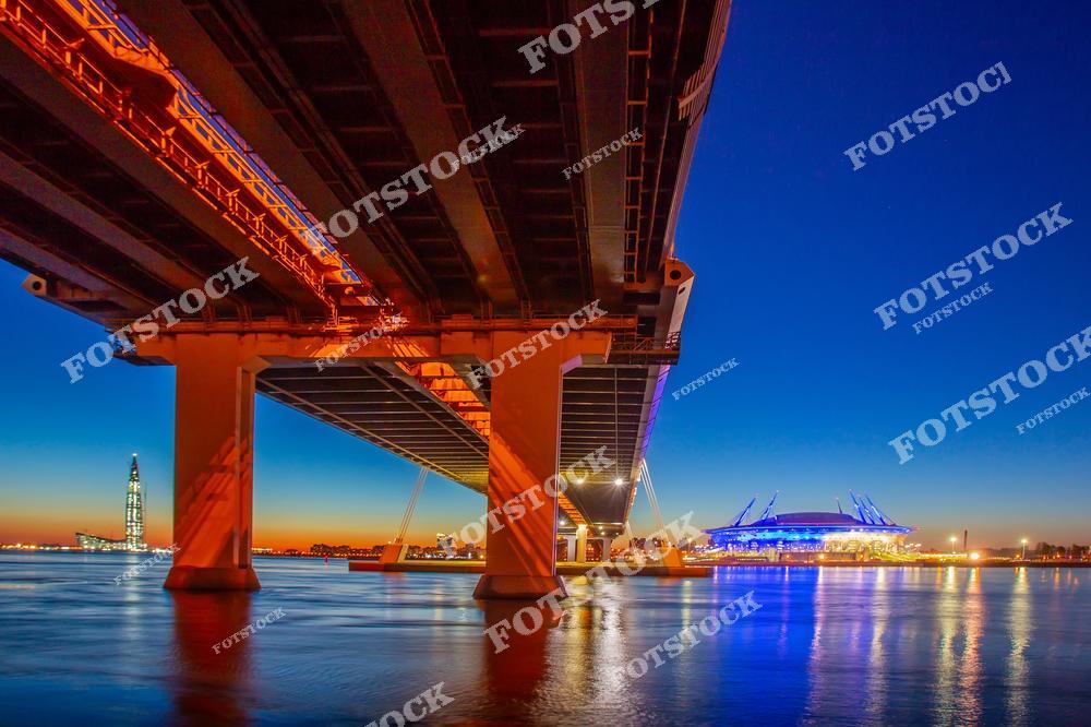 Фотография панорамного вида Санкт-Петербурга с высоты, охватывающая Васильевский остров, Неву и Финский залив, а также городскую набережную и магистраль. Снимок передает атмосферу белых ночей в северной столице России: мягкий свет вечера отражается в воде и на фасадах зданий, создавая романтическую и живописную сцену. Панорамный ракурс позволяет увидеть масштаб города, исторические и современные сооружения, а также динамику городских улиц и магистралей. Изображение идеально подходит для туристических публикаций, постеров, сувенирной продукции и коллекций фотографий Санкт-Петербурга.